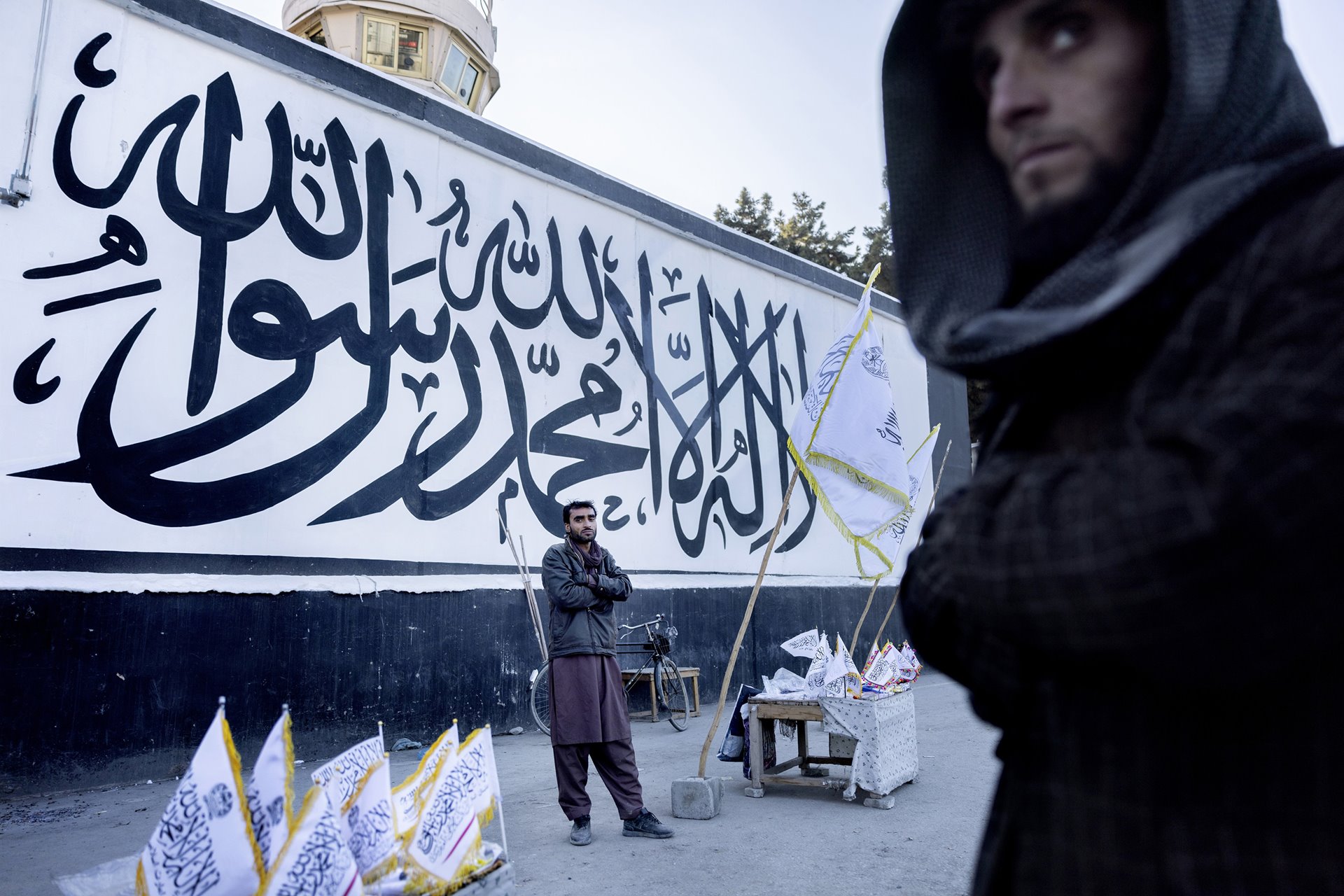The Islamic declaration of faith, &quot;There is no God but Allah, and Muhammad is His messenger&quot;, covers the wall of the former US Embassy in Kabul, Afghanistan. In front of the wall, street vendors sell Taliban flags and other merchandise. US diplomats negotiated with the Taliban in August 2021 to spare the embassy after the evacuation &nbsp;in exchange for future aid.