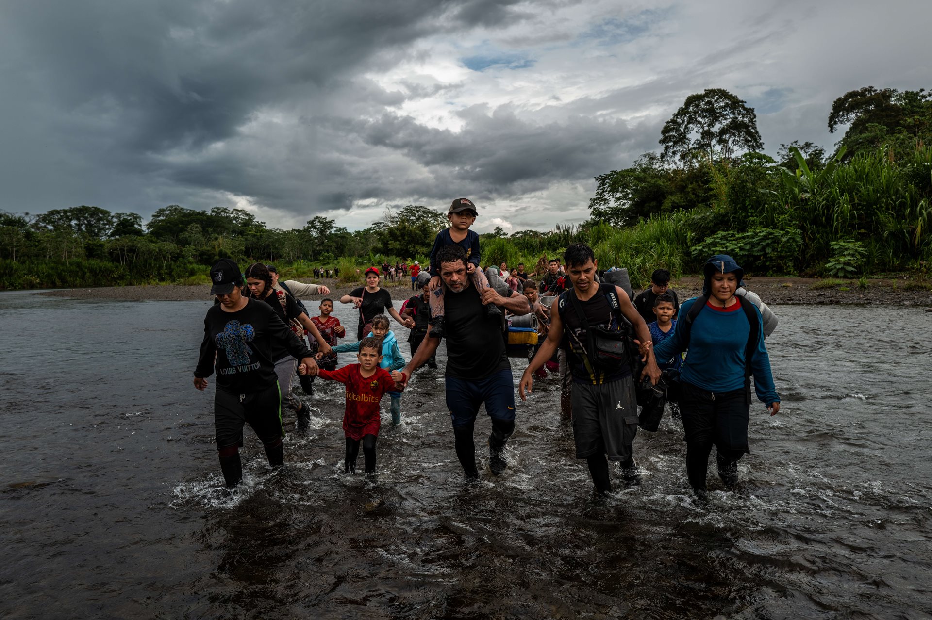 Several groups of migrants cross the Tuquesa River. Juan Carlos Taborda (47, center) carries his three-year-old grandson, also named Juan Carlos Taborda, during the final stretch of their trek through the jungle. They are at Come Gallina, the last migrant camp in the jungle before reaching the Indigenous community of Bajo Chiquito in Panamá.
