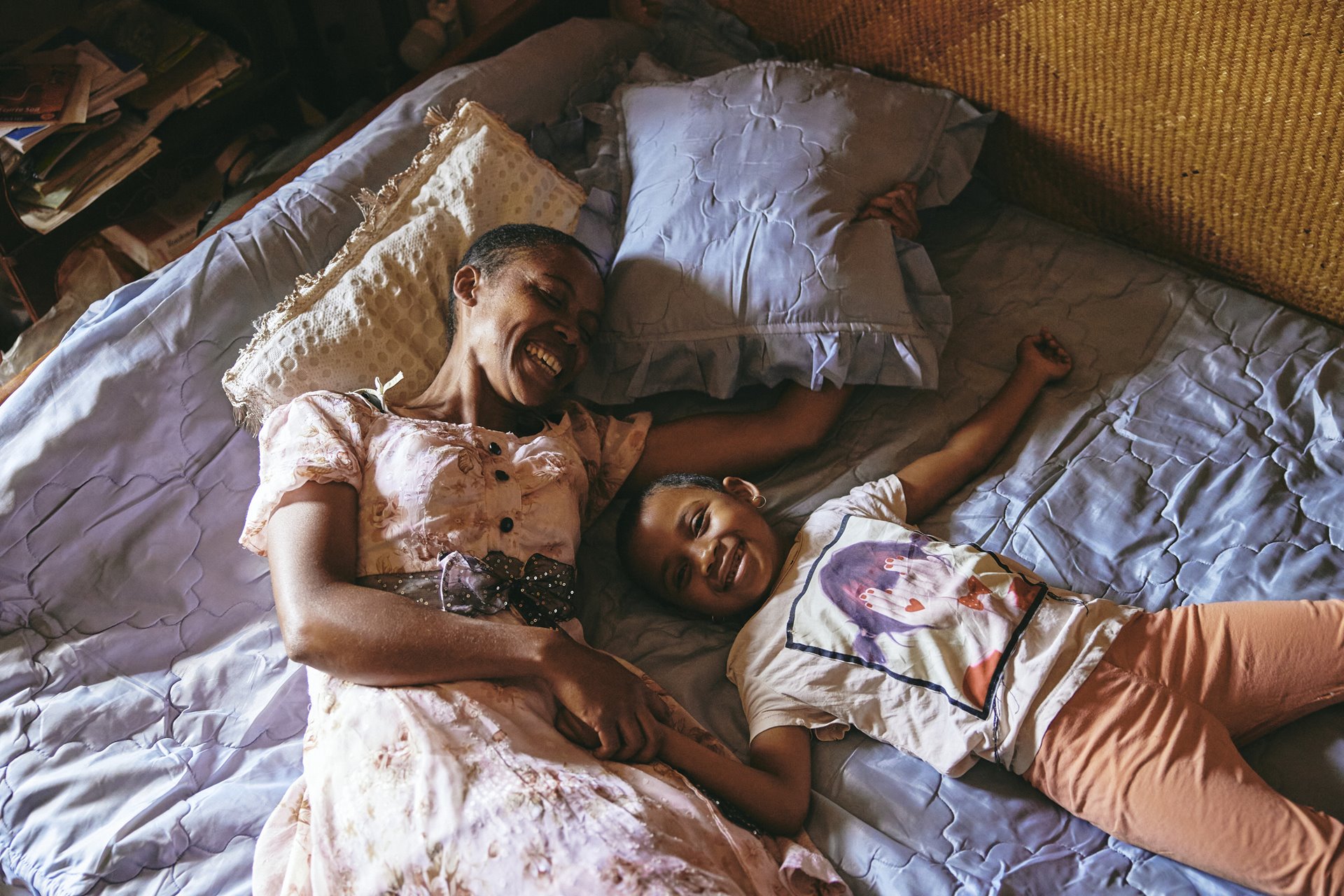 Fara and her daughter Odliatemix lie together on the bed they share with Dada Paul, Fara&rsquo;s father, in Antananarivo, Madagascar. Fara is the sole provider for the family of three and struggles to balance her time between work, caring for her father, and spending time with her daughter.
