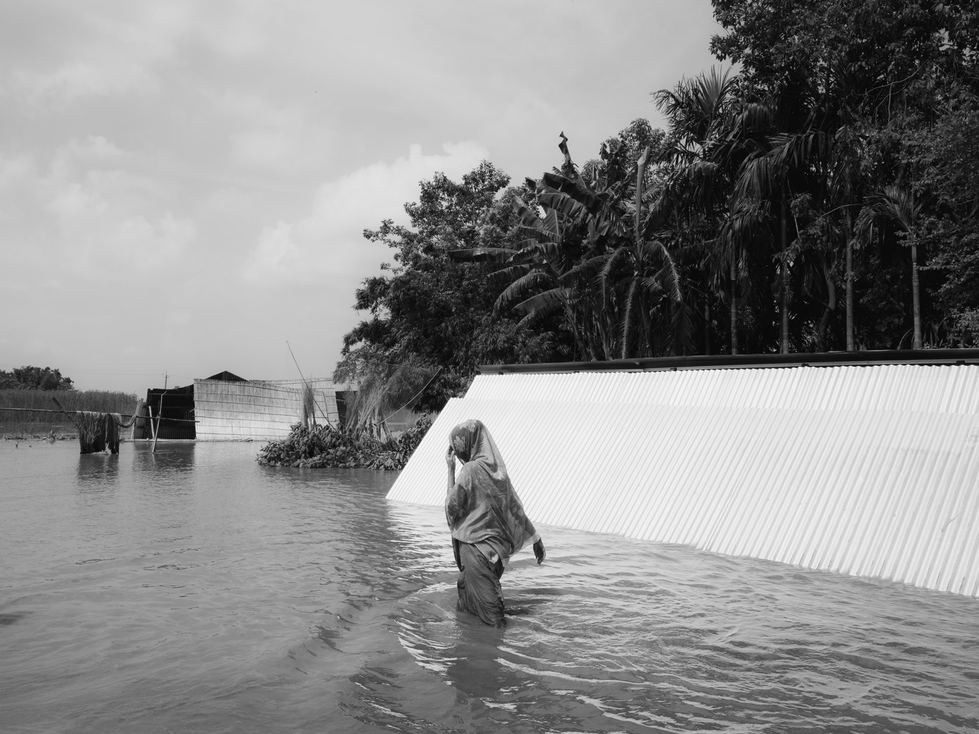 In Kharbali, a Bengali Muslim woman navigates floodwaters during Assam&#39;s five-month monsoon season. In July 2023, devastating floods affected lower Assam, a region that is predominantly Bengali Muslim. Approximately 35,000 sought refuge in flood relief camps. The floods obliterate homes, farms, and crops, intensifying struggles for National Register of Citizens (NRC) inclusion amidst political and environmental precarity. Jania, Barpeta, Lower Assam, India.