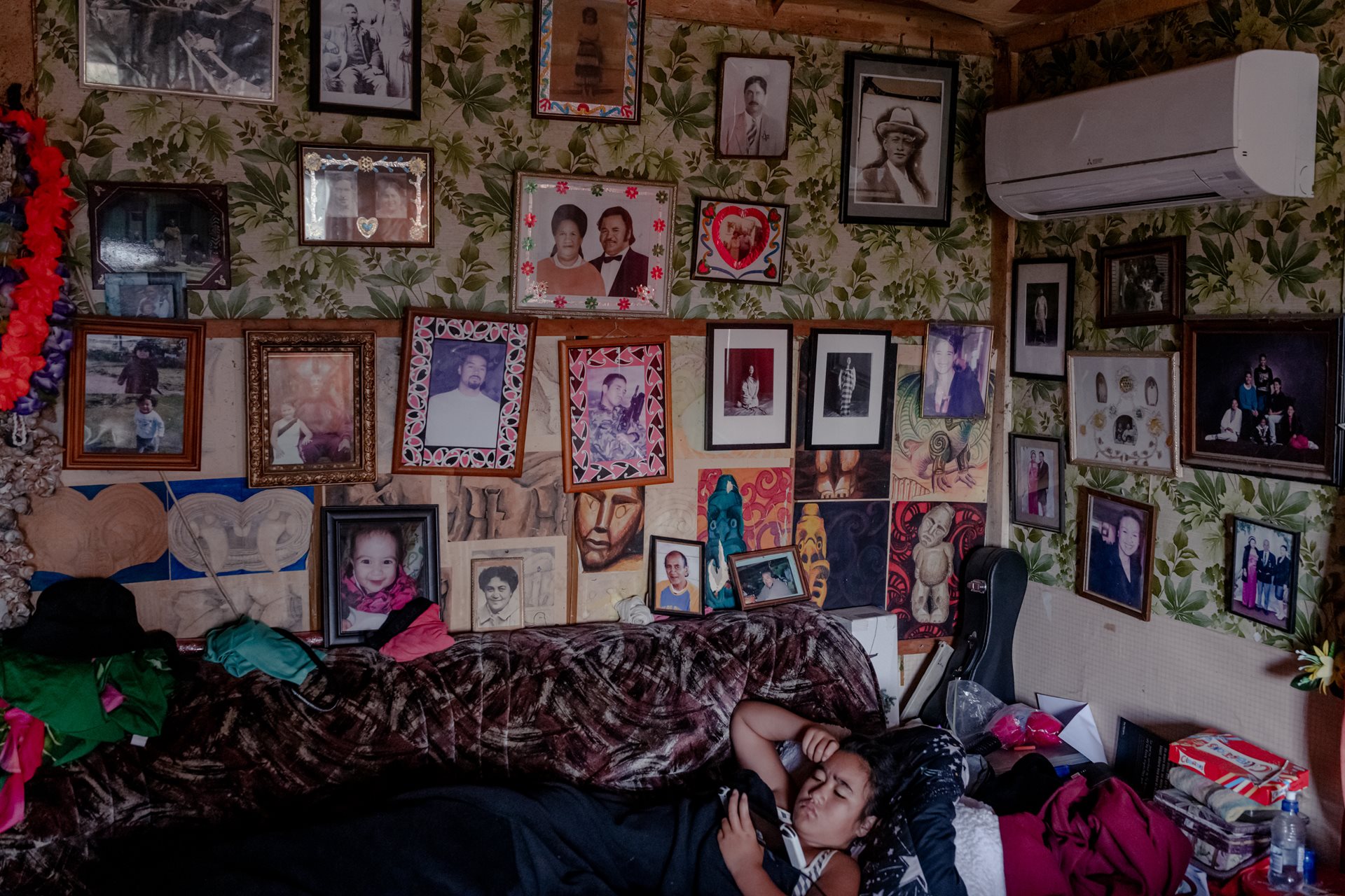 Ruiha Te Tana (12) relaxes at her grandfather&#39;s home in Ruatoki, New Zealand. Built by an ancestor in 1916, the homestead serves as a living archive of Tūhoe history. The wall of photographs visually represents Te Tana&#39;s whakapapa, or genealogy, a cornerstone of Maori culture. Whakapapa connects individuals to their ancestors, land, and tribal identity, providing a sense of belonging and continuity.