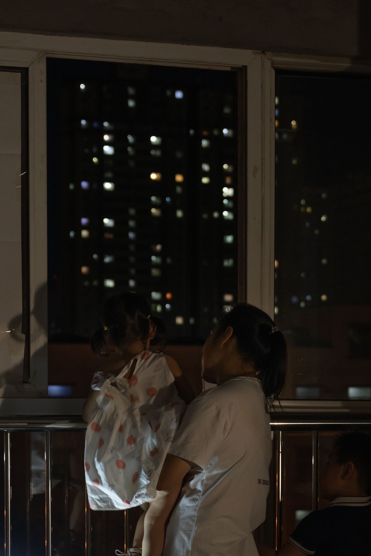 A mother and child gaze out at city lights in Xi&rsquo;an, China. In contrast, their own home is lit from below with a solar powered lamp due to lack of electricity in their building.