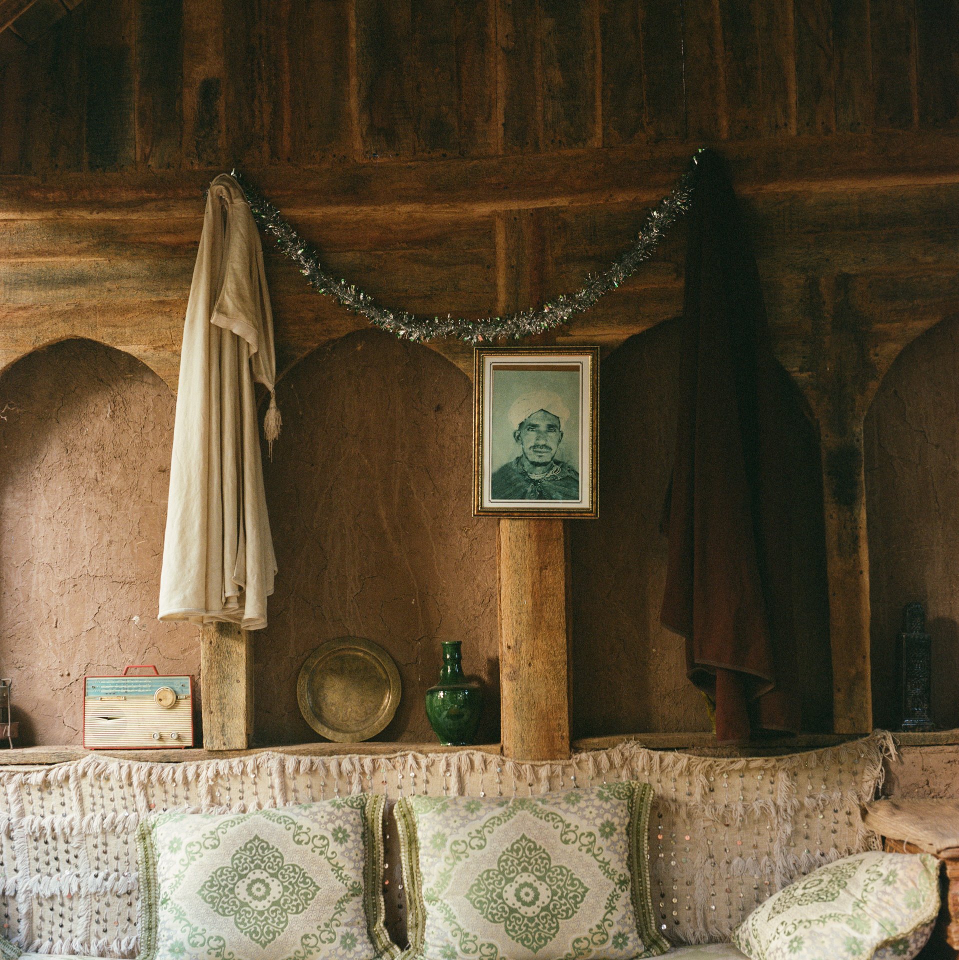 <p>This domestic interior tableau features djellabas (robes), a traditional tea tray, and an ancestral portrait alongside a radio (formerly the main source of contact with the world across the dunes) in Zagora, beside the Oued Draa river in eastern Morocco.</p>
