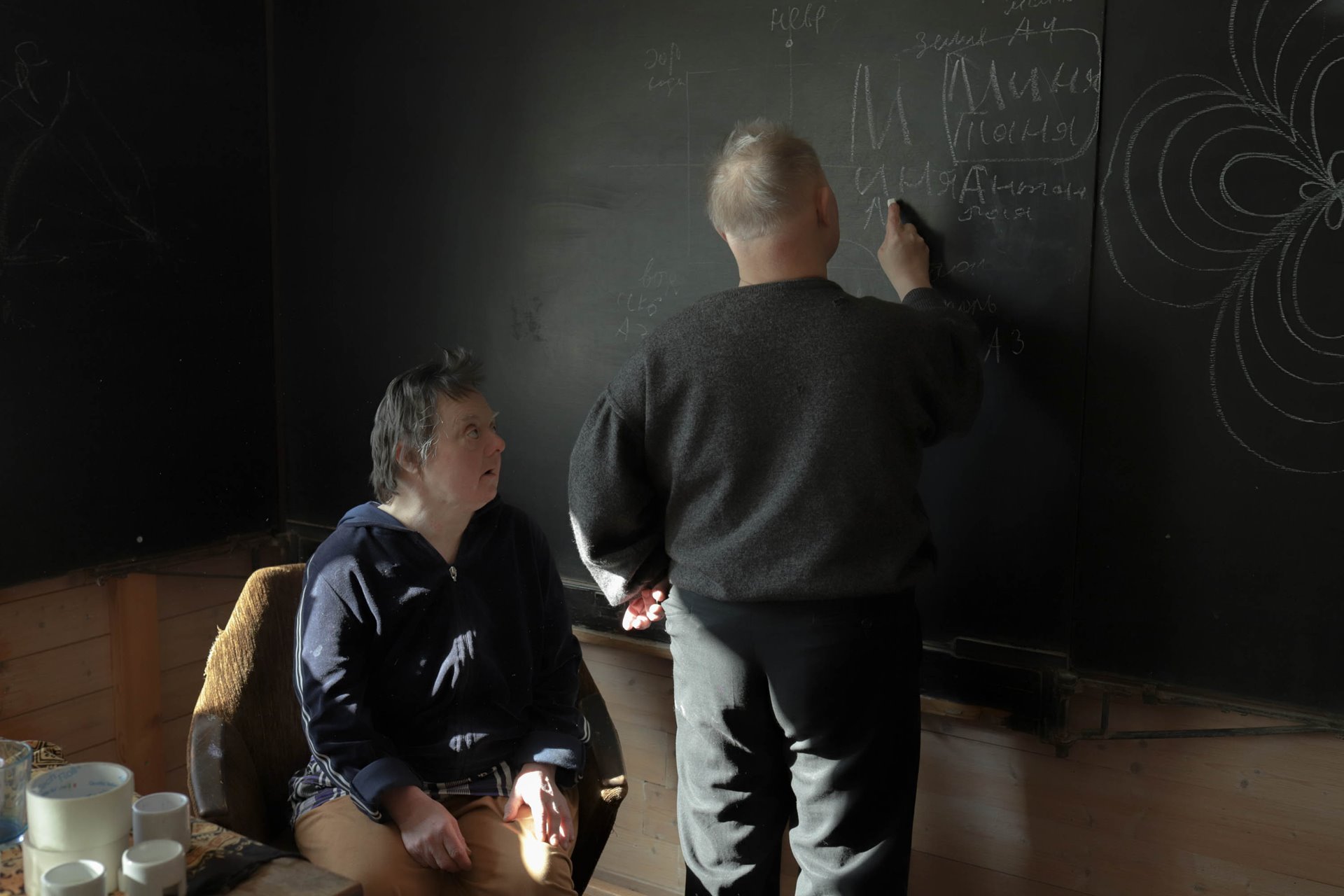 Minya writes on the blackboard in the village study room, while Tatyana looks on.