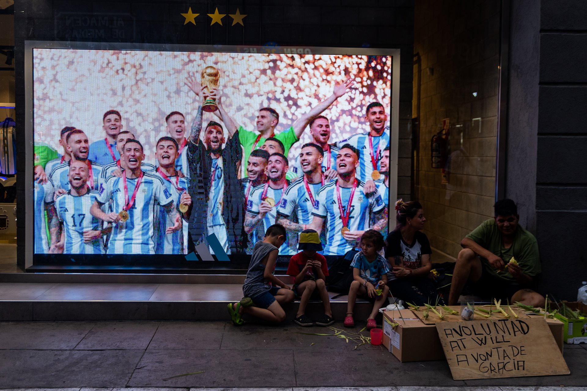 A family sells handicrafts to tourists next to a storefront display of Lionel Messi and his teammates lifting the World Cup in Buenos Aires, Argentina. Inflation and poverty are at record highs in Argentina, where annual inflation has hit nearly 100% and more than 50% of children live in poverty.&nbsp;