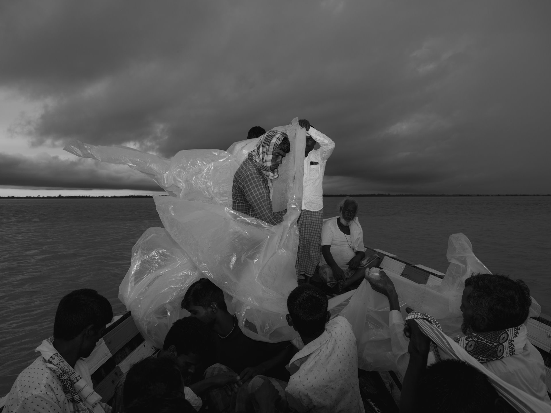 Bengali Muslim villagers, accustomed to the yearly floods, are ferried by community boats to the mainland from tiny chars, sandbar islands formed by silt deposits on the Brahmaputra. Living and farming on chars, these villagers face social, political, and economic marginalization. As monsoon clouds loom in the distance, they seek refuge under plastic sheets, embodying resilience amid their struggle for identity and livelihoods along the Brahmaputra River. Uttar Lakhipur, Sontoli, Kamrup/Barpeta District, Lower Assam, India.