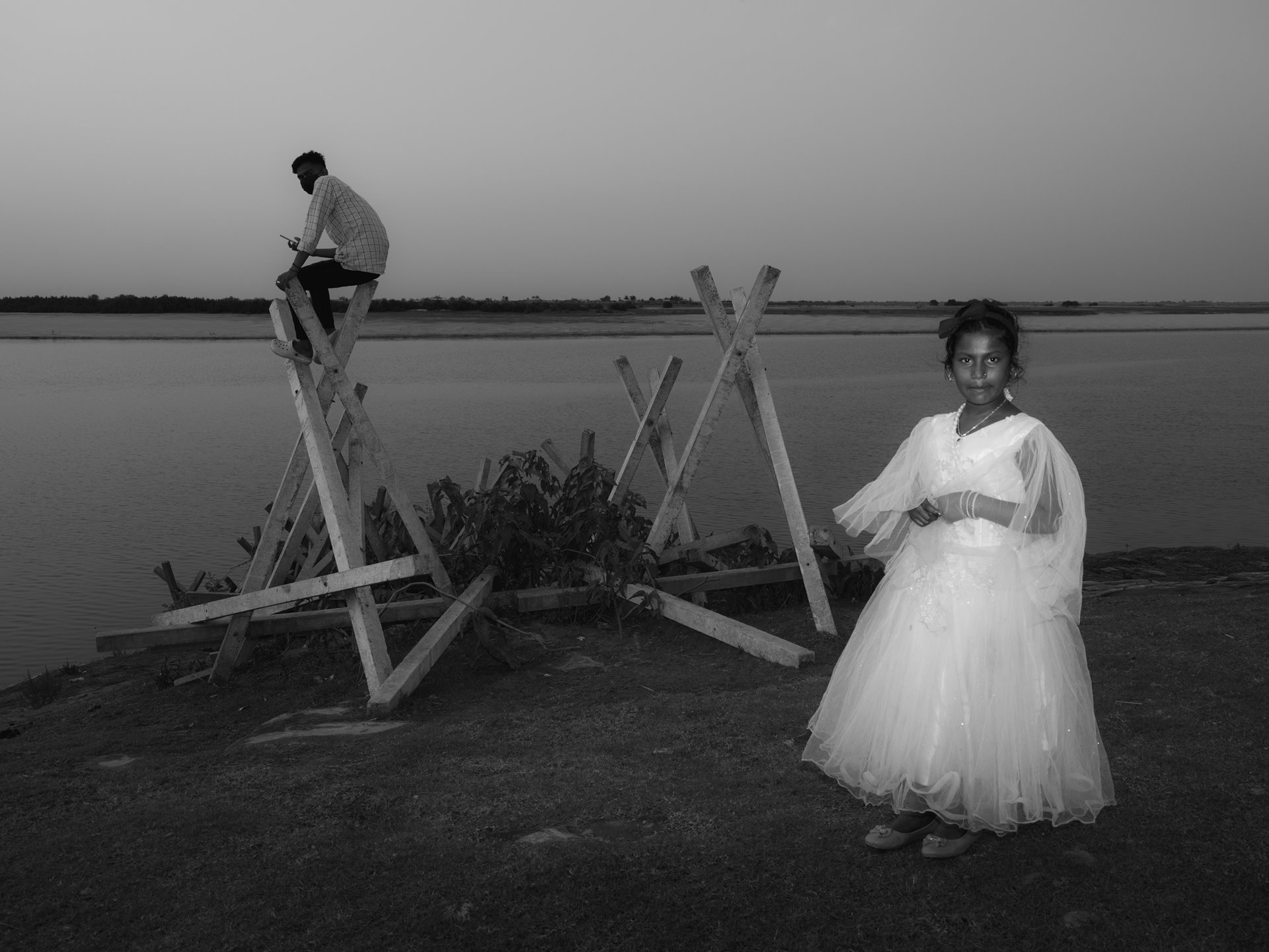 A young Bengali Muslim boy attempts to secure a network signal for his cellphone atop a concrete &quot;porcupine&quot; while a girl celebrates Eid in her finest festive attire. The &quot;porcupine&quot; is a flood precaution introduced by the state&rsquo;s Brahmaputra Board but failed to match the effectiveness of traditional methods used by Indigenous Assamese. In the eyes of local communities, the concrete structures have become a symbol of the incompetence and corruption of the board. Panikhaiti, Shontuli, Kamrup District, Lower Assam, India.