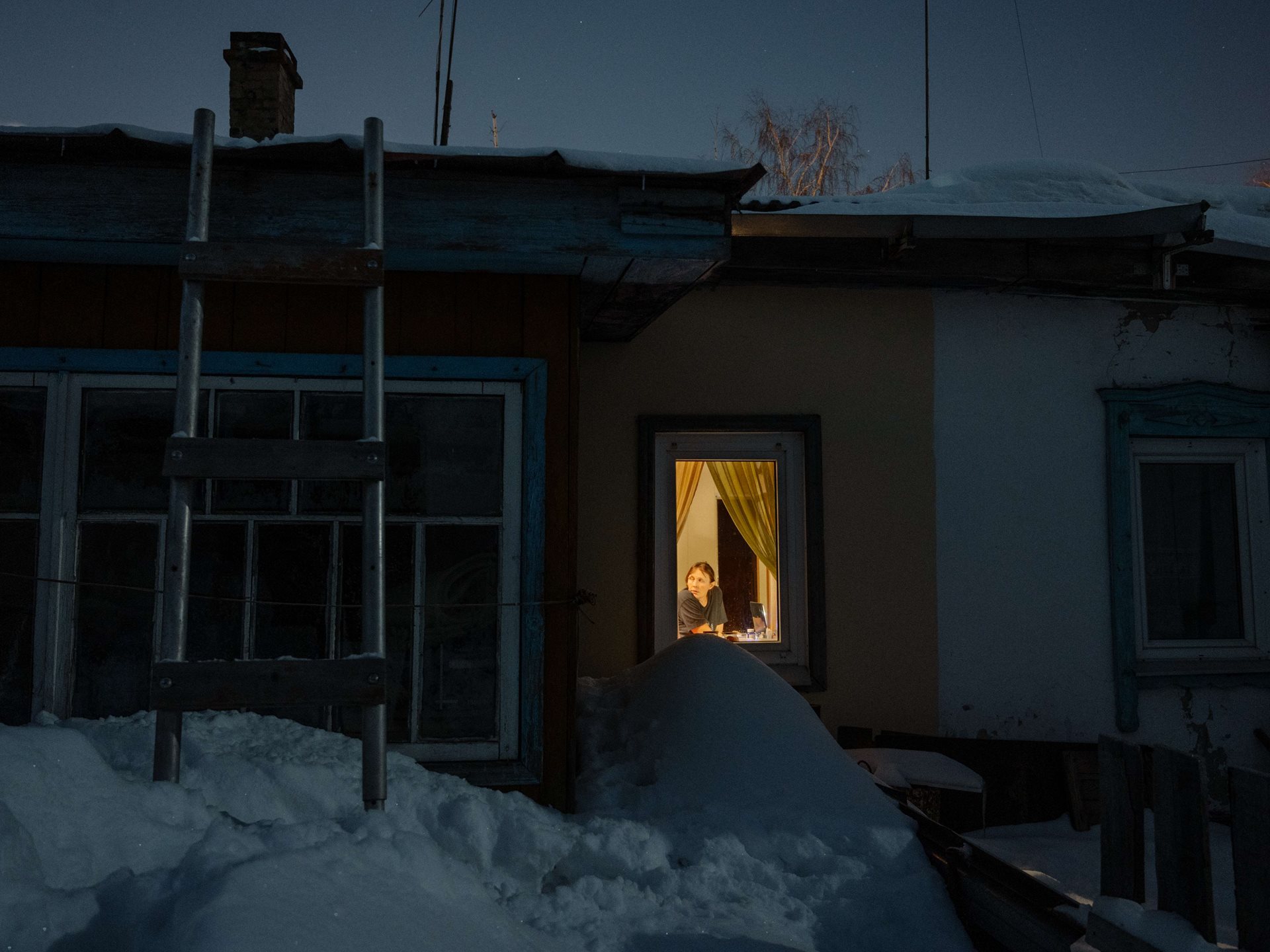 Aliona&rsquo;s mother looks out of a window at home, in Tomsk, Russia. At the end of winter, there is so much snow in Tomsk that it starts to reach the windowsill and seems to be squeezing the walls.