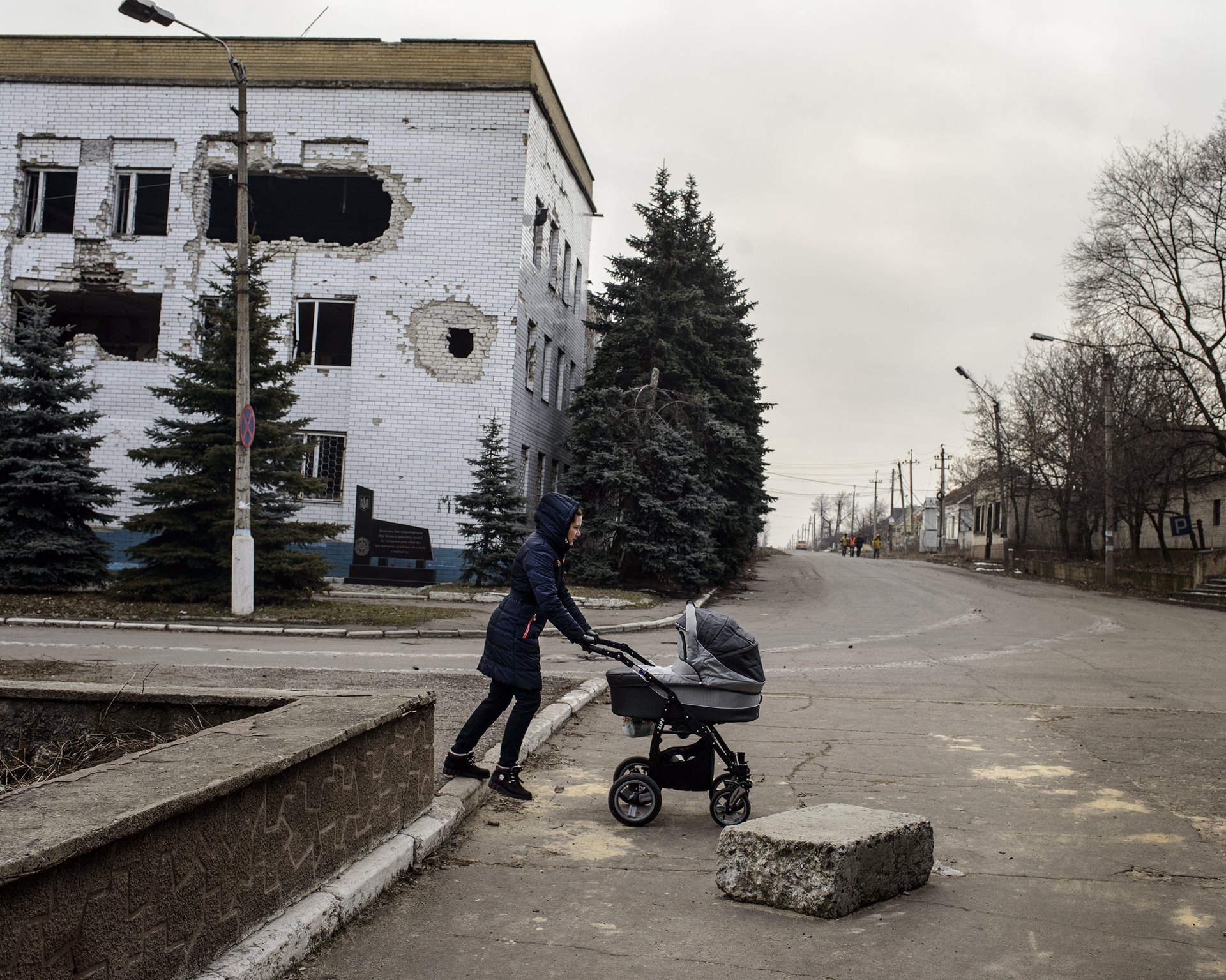 A woman pushes a baby carriage across Lenin Street, past a police station destroyed during fighting in 2014, in Marinka, Donbas, Ukraine.