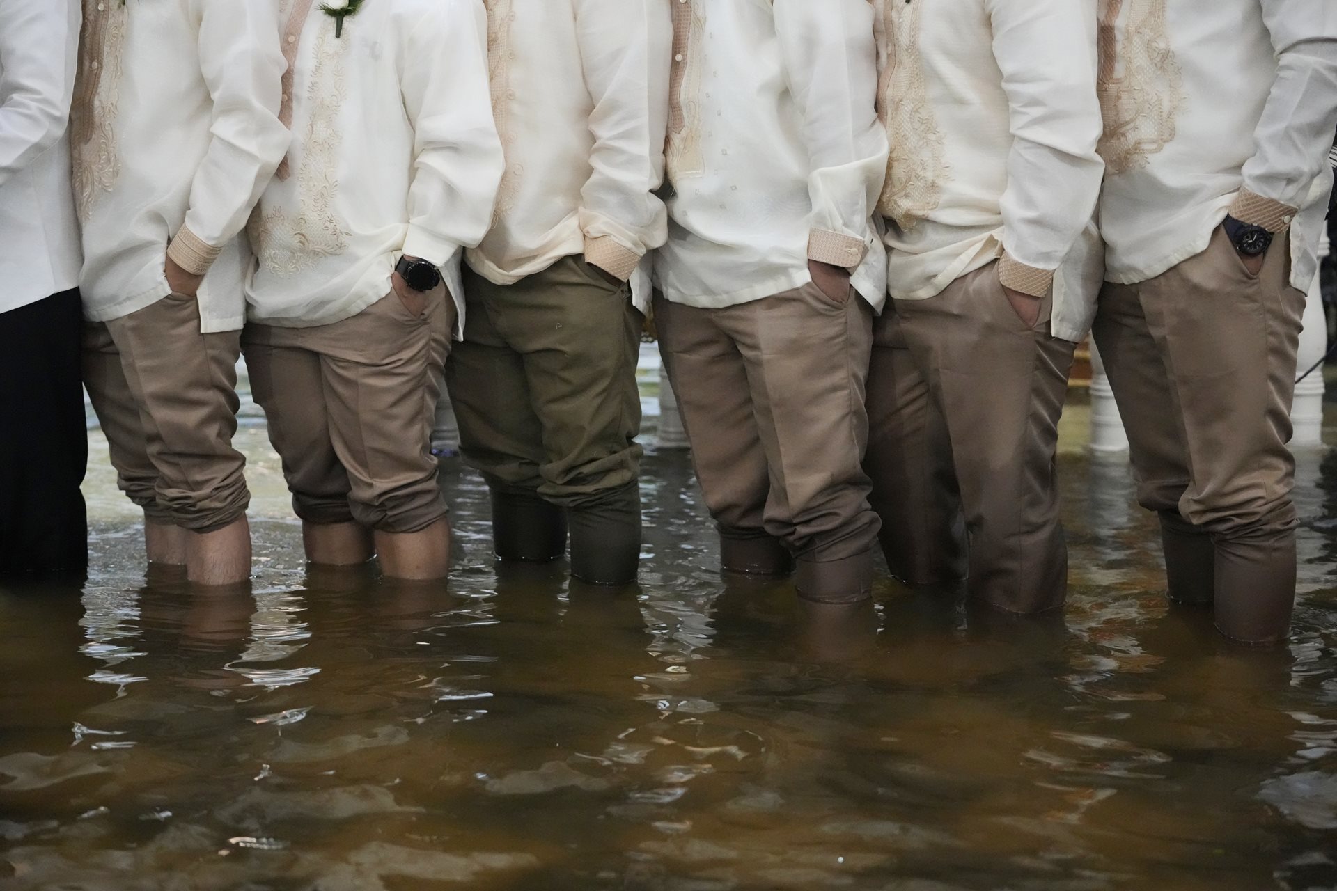Groomsmen and guests wearing barong tagalog, the national dress of the Philippines, stand knee-deep in floodwaters. Local advocates increasingly link flooding to systemic failures in infrastructure and resource management. Malolos, Bulacan province, Philippines