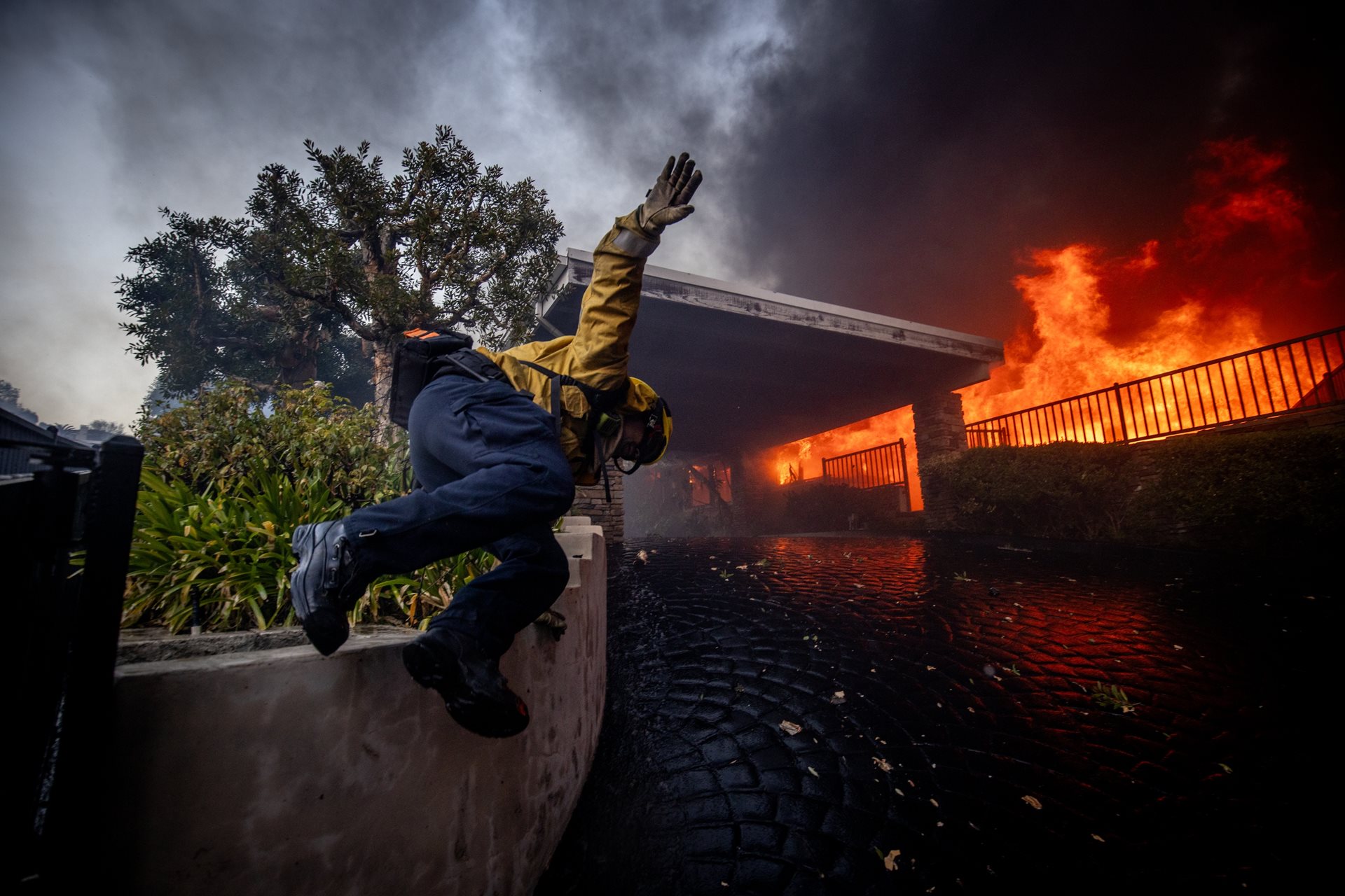 <p>A firefighter jumps over a fence while battling the Palisades Fire. More than 7,500 emergency personnel, including incarcerated firefighters and the National Guard, were deployed to combat the rapidly spreading blazes. Los Angeles, California, United States</p>
