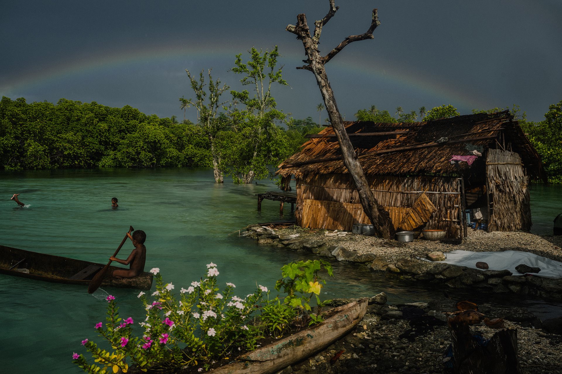 After a severe storm, children play in canoes among houses surrounded by the encroaching ocean. For the younger generation, daily life is defined by the constant threat of the disappearance of their land. Fanalei Village, Fanalei Island, South Malaita, Solomon Islands.