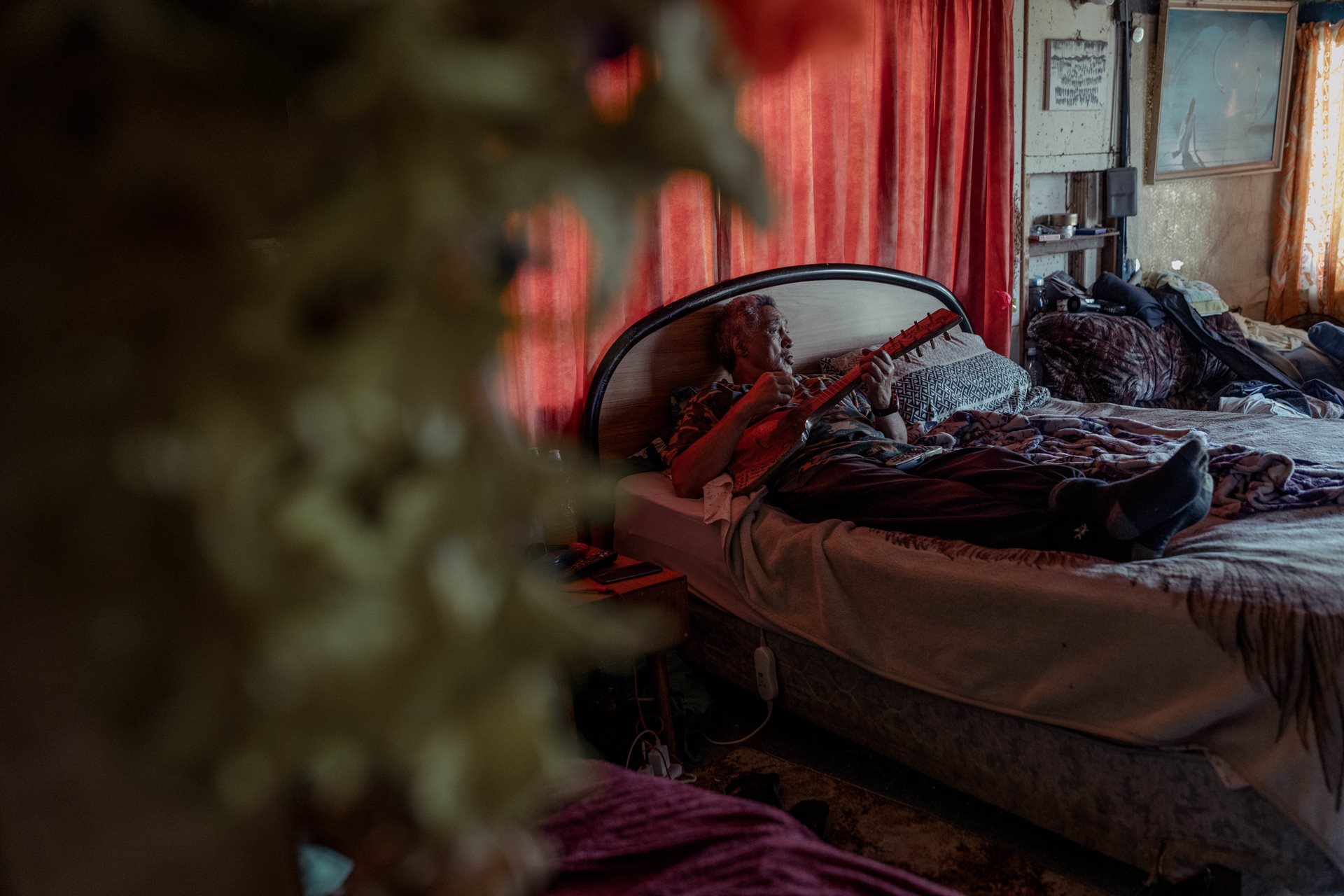 John Rangikapua Teepa (68) plays the ukulele in his bedroom at home, in Ruatoki, New Zealand. He suffers from poor health and spends most of his time resting, while trying to help on the farm very early in the day. Like many Tūhoe, John Teepa spent a few decades living in the city, away from his ancestral land.