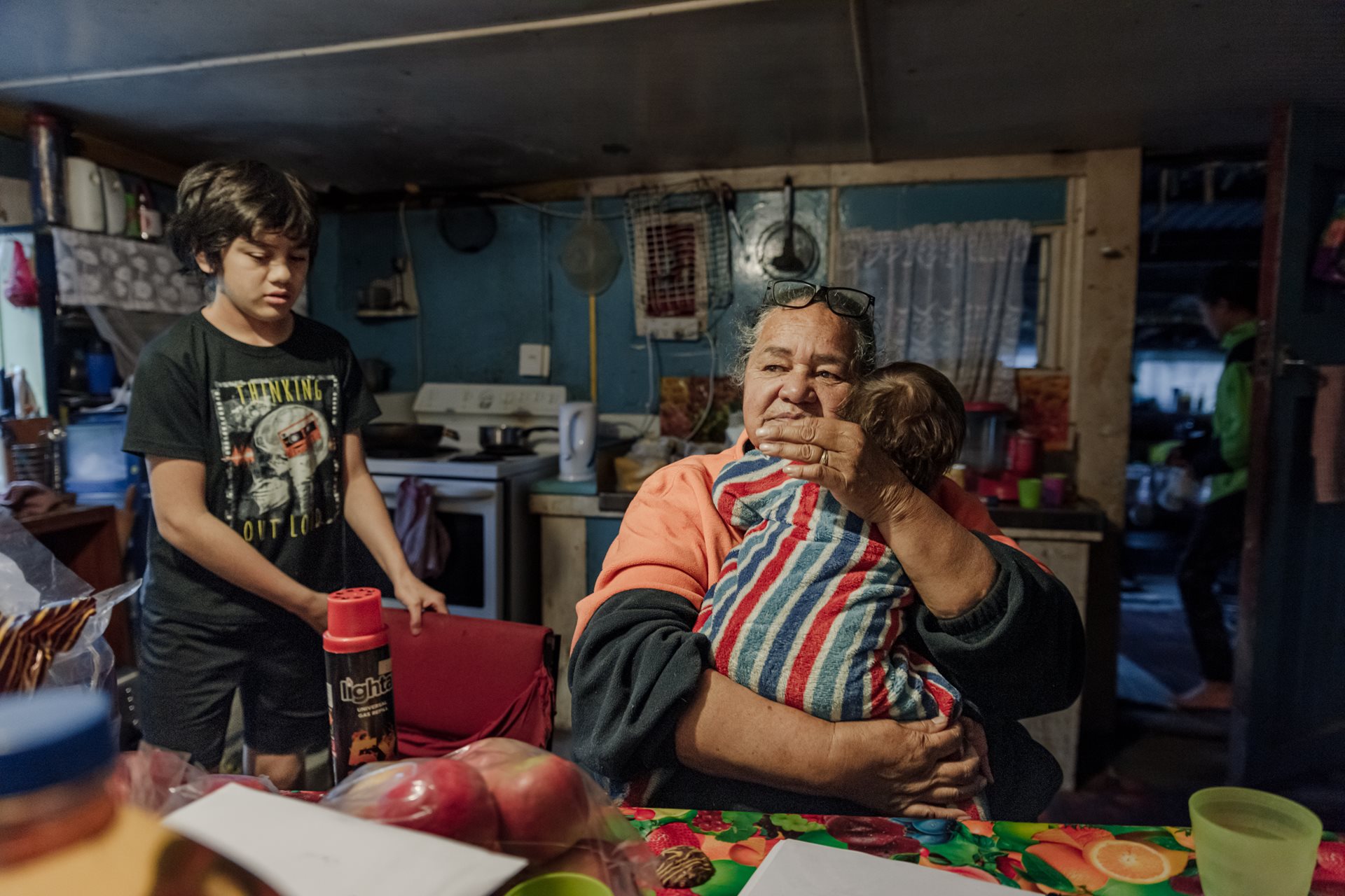 Carol Teepa (64) sits in her kitchen in Ruatoki, New Zealand, with her youngest grandchild, Mia, and her whāngai son, Wanea, one of more than 20 children she adopted according to Māori whāngai custom, to strengthen traditions and family ties. Whāngai children often maintain relationships with both birth and adoptive families. This open arrangement helps preserve cultural heritage and strengthens family ties.