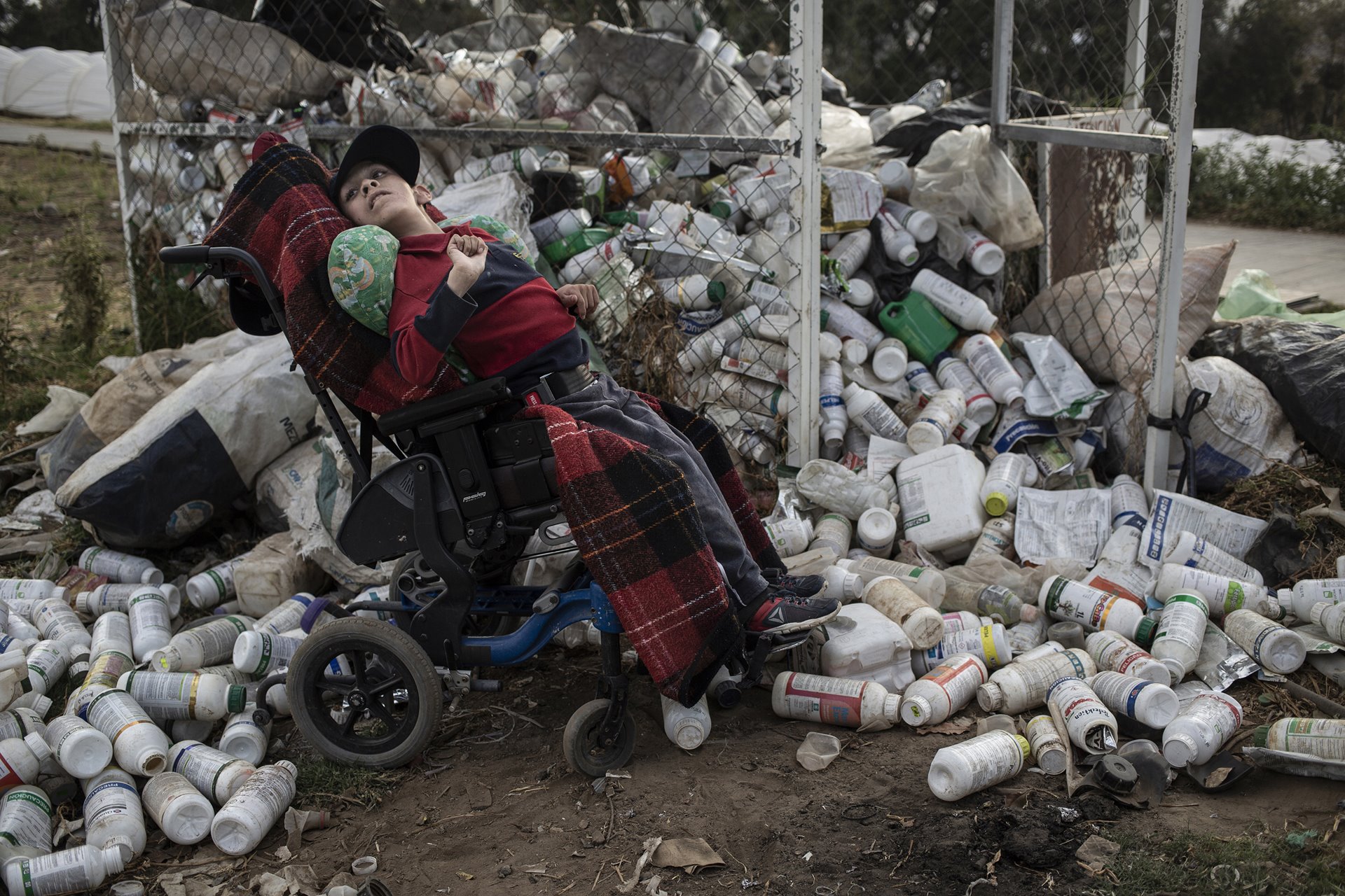 Sebastián (18), who lives with hydrocephalus (a neurological disorder caused by a build-up of fluids in cavities within the brain), waits in front of a garbage collection point for empty agrochemical containers, while on a walk near home with his father, in Villa Guerrero, Mexico. Such collection cages are also in use in other parts of the world. Authorities in the US and Australia recommend triple-rinsing of containers before disposal in this manner.