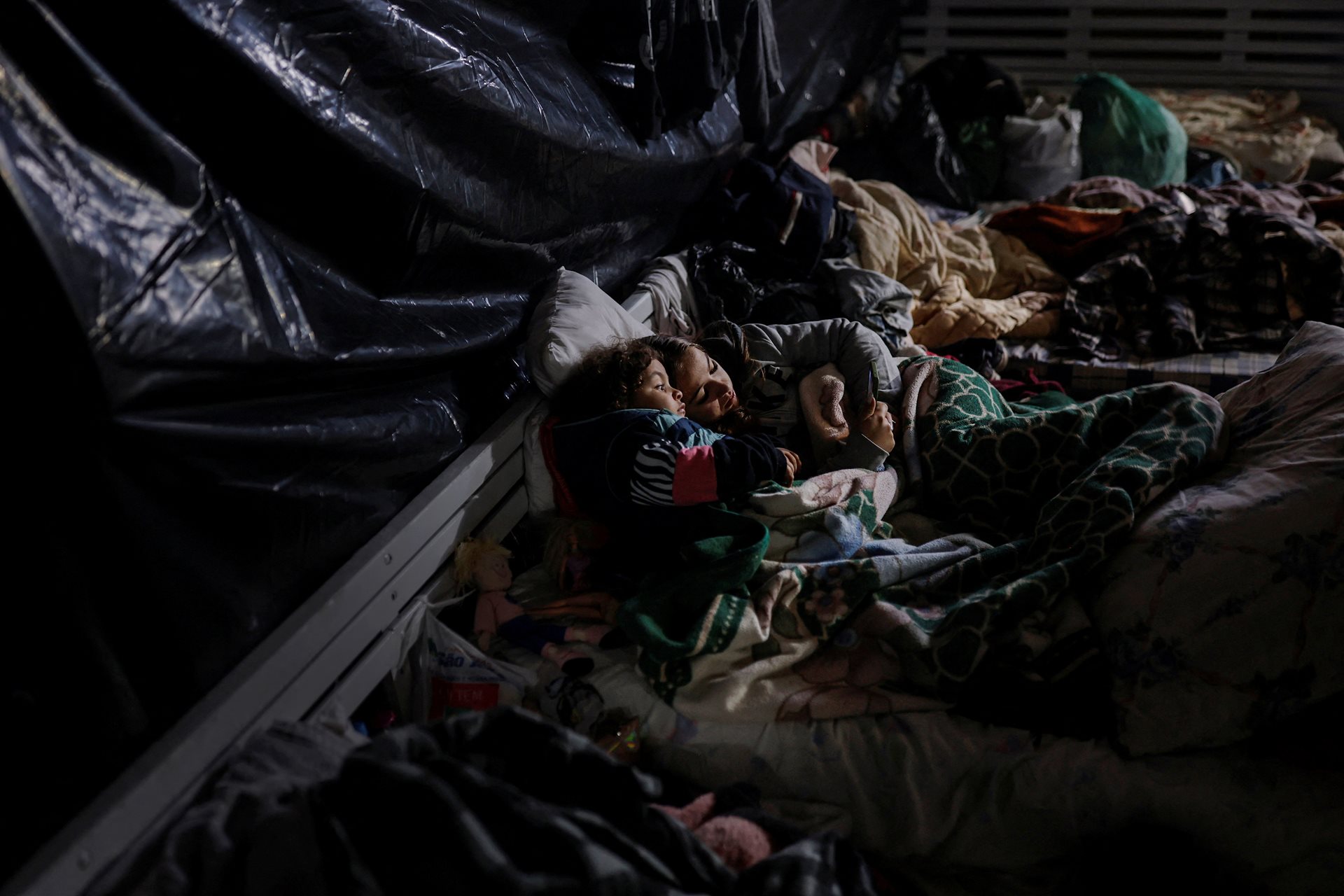 Two girls watch cartoons on a phone at a truck factory warehouse, after being displaced by floods. Washed-out roads have led to food and water shortages, disrupting the lives of over two million people. Eldorado do Sul, Rio Grande do Sul, Brazil.