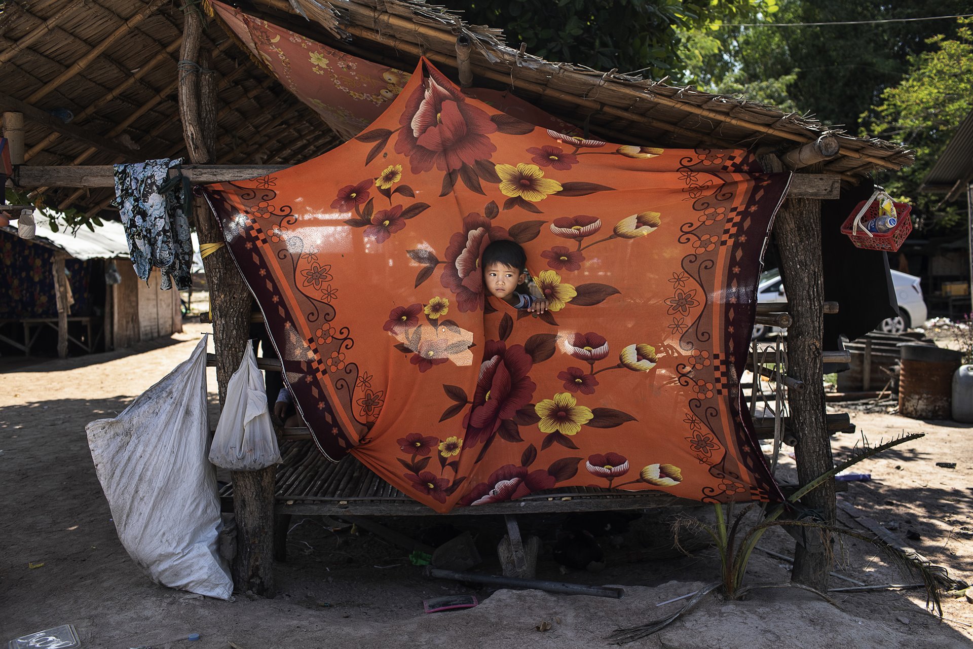 Korng (3), Ry Ly&rsquo;s son, peeks through a hole in a sheet that shades the family&rsquo;s yard from the sun. Ry Ly is a garment factory worker who also has a seven-year-old daughter with her husband, who remains supportive of her. Kampong Speu, Cambodia.